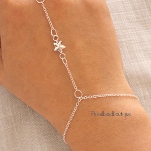 Può includere: Una delicata catena da mano argentata con un ciondolo a forma di stella e un anello di collegamento. La catena si estende dall'anulare al polso, con un piccolo pendente a forma di stella. Gioiello su sfondo chiaro.