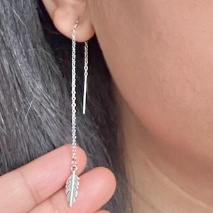 以下が含まれることがあります： 銀色のチェーンイヤリング。片方のチェーンには羽根のチャーム、もう片方のチェーンにはストレートな銀のバーがぶら下がっています。画像の左下にはFloralbeadboutiqueと書いてあります。
