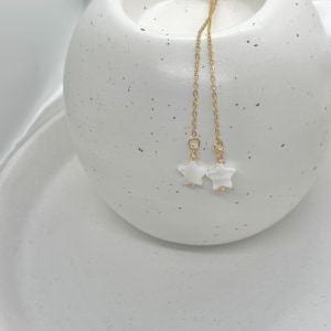 Puede incluir: Un par de pendientes dorados con colgantes blancos en forma de estrella. Los pendientes cuelgan de finas cadenas doradas, con las estrellas sobre un fondo de cerámica blanca moteada. Un accesorio sencillo y elegante.