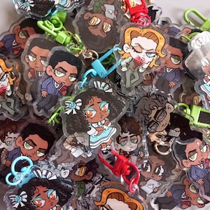 May include: A selection of cartoon character keyrings with diverse designs. The keyrings depict characters with varied hairstyles, clothing, and expressions. Each keyring has a colourful clasp, with shades of red, green, and blue.