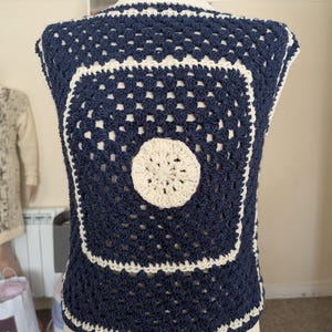 Puede incluir: Un top sin mangas de ganchillo con un patrón de granny square en azul marino oscuro y un motivo circular central de color crema. La prenda está rematada con un borde de ganchillo color crema, mostrando un diseño geométrico calado.