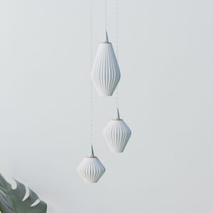 Może przedstawiać: Trzy białe lampy wiszące o żebrowanym, geometrycznym wzorze zwisają z sufitu. Każda lampa ma kształt łzy z miedzianą górą. Lampy zawieszone są na gładkim, białym tle, tworząc minimalistyczną estetykę.