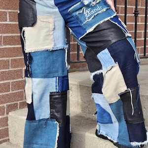 Könnte beinhalten: Weite Patchwork-Jeans mit ausgefransten Kanten und einem schwarzen Lederpanel. Die Jeans sind aus verschiedenen Blautönen von Denim gefertigt und haben einen beigefarbenen Patch mit dem aufgestickten Wort "Wandr".