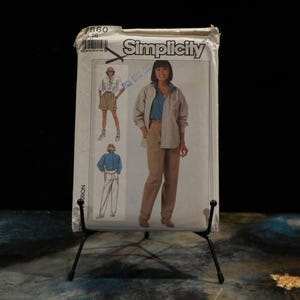 Pode incluir: Um pacote de padrões de costura Simplicity vintage, com ilustrações de designs de vestuário. O pacote mostra uma mulher com camisa e calças bege, e outros designs. O pacote é suportado por um suporte de metal preto.