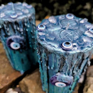 Könnte beinhalten: Zwei tealblaue Kerzen mit schwarzen Wachstropfen. Jede Kerze hat ein Pentagramm-Design und ist mit mehreren blauen bösen Augen-Anhängern und klaren Quarzkristallen verziert.