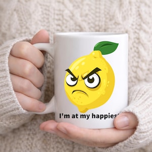 May include: White ceramic mug featuring a cartoon lemon with an angry expression and a green leaf. The mug has the text "I'm at my happiest" printed on it. The mug is held by a person wearing a beige sweater.
