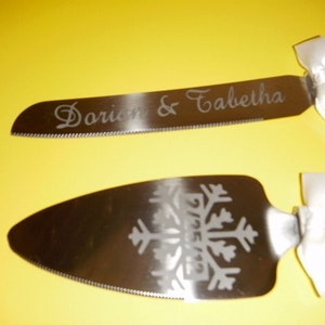 May include: Two silver cake knives with white satin bows. One knife has the names "Dorien & Tabitha" engraved on the blade. The other knife has a snowflake design engraved on the blade.