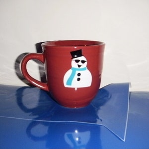 May include: A red ceramic mug with a snowman design. The snowman is wearing a black top hat, sunglasses, and a turquoise scarf.