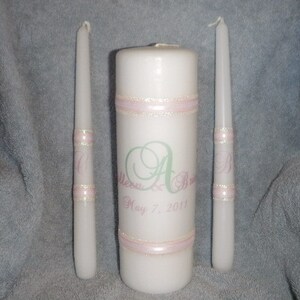 May include: Two white taper candles with pink and gold ribbon accents and a larger white pillar candle with pink ribbon accents. The pillar candle has the text "Melissa & Andrew May 7, 2011" printed on it.