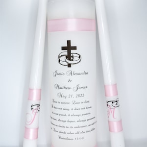 May include: Three white unity candles with pink ribbon accents. The center candle has a cross and two rings, and the text "Jamie Alexandra & Matthew James May 21, 2022" and a quote about love from Corinthians 13:4-8.