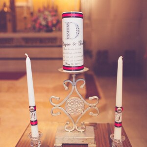 Puede incluir: Dos velas blancas con detalles de cinta negra y rosa en candelabros de plata. Una vela blanca más grande con detalles de cinta negra y rosa y el texto "Megan & Sean" y "May 10, 2014" está en un soporte decorativo de plata.