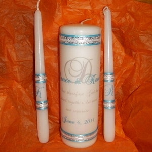 May include: Two white taper candles and a larger white pillar candle with blue and silver ribbon accents. The pillar candle has the text "Renee & Kevin" and "June 4, 2011" on it.