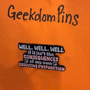 May include: Black enamel pin with the text "WELL, WELL, WELL if it isn't the CONSEQUENCES of my own EXECUTIVE DYSFUNCTION" in white and pink. The pin is set against an orange background with the words "Geekdom Pins" in black.