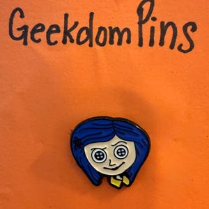 May include: A metal enamel pin featuring a cartoon character with blue hair and button eyes, positioned below the handwritten text Geekdom Pins. The character has a pale face, a small black flower in the hair, and wears a yellow collared shirt.