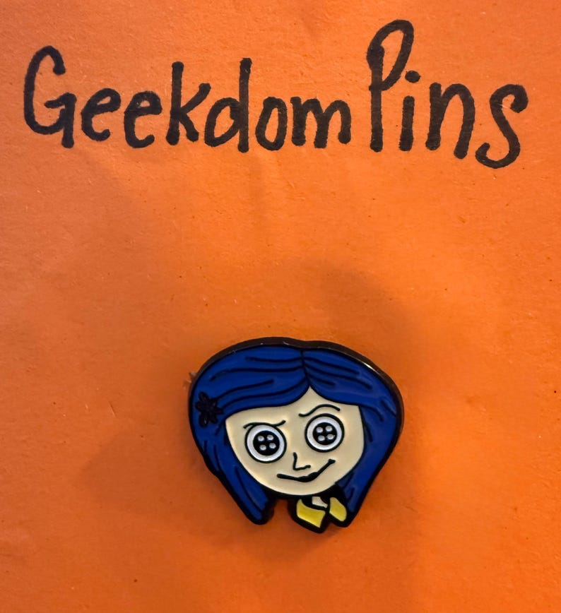 May include: A metal enamel pin featuring a cartoon character with blue hair and button eyes, positioned below the handwritten text Geekdom Pins. The character has a pale face, a small black flower in the hair, and wears a yellow collared shirt.