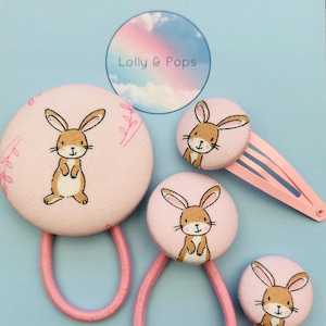 May include: A set of three pink hair accessories featuring a brown bunny rabbit design. The set includes two hair scrunchies and one hair clip.