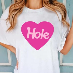 Puede incluir: Camiseta blanca con un gran corazón rosa. La palabra "Hole" está impresa en el centro del corazón. La camiseta tiene mangas cortas y un corte holgado.