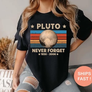 May include: Black t-shirt with a Pluto graphic and the text "PLUTO NEVER FORGET 1930-2006". The design uses a retro colour scheme with blue, orange, and brown stripes. This is a casual wear item.