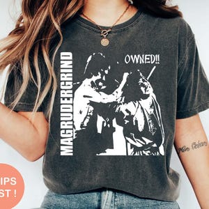 May include: Dark gray t-shirt with a white graphic featuring two figures and the text "MAGRUDERGRIND" and "OWNED!!". The shirt has a vintage, worn look.