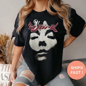 May include: Black t-shirt featuring a face graphic and the name "Bjork" in burgundy. The shirt has short sleeves and a crew neck. The graphic is a stylized portrait. The shirt is worn by a person.