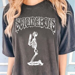 May include: Dark grey t-shirt featuring the text "SUICIDEBOYS" in a distressed font. A white skeleton is kneeling in prayer. The shirt has a relaxed fit, suitable for casual wear.