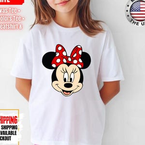 May include: White t-shirt featuring a Minnie Mouse graphic with a red bow with white polka dots. The shirt has the text "BELLA-CANVAS 3001Y WHITE" and "MADE IN THE USA" on it. The shirt is paired with pink shorts.