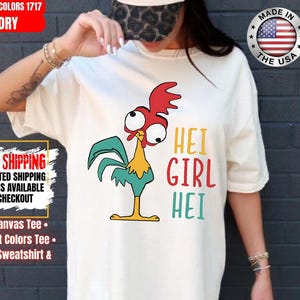 May include: Ivory Comfort Colors t-shirt with a cartoon rooster and the words "HEI GIRL HEI". The shirt also has the text "Made in the USA" and "Fast Shipping" on it. The t-shirt is made of ivory-colored fabric.