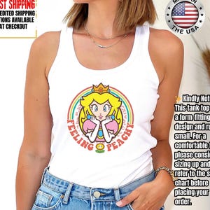 May include: White tank top featuring a cartoon princess design with the text "Feeling Peachy" in a rainbow circle. The princess has yellow hair, a gold crown, and pink accents. Made in the USA.