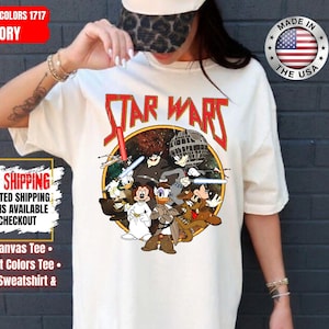 Op de afbeelding: Ivoor kleurig Star Wars grafisch T-shirt met een cartoon mix van Star Wars en Disney karakters. Het ontwerp bevat het Star Wars logo in rood, een Death Star en diverse karakters. Het shirt is een Comfort Colors 1717 Bella-Canvas Tee.