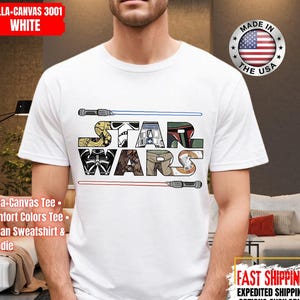 May include: White t-shirt with the words "STAR WARS" in a block-letter design, incorporating Star Wars imagery. Blue and red lightsabers are above and below the text. The shirt is made in the USA.