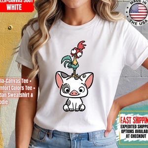 K&ouml;nnte beinhalten: Wei&szlig;es Bella+Canvas 3001 T-Shirt mit einem Cartoon-Schwein und Hahn-Design. Das Schwein ist wei&szlig; mit schwarzen Flecken und rosa Ohren, und der Hahn ist gr&uuml;n, rot und gelb. Das Shirt hat auch den Text "MADE IN THE USA".