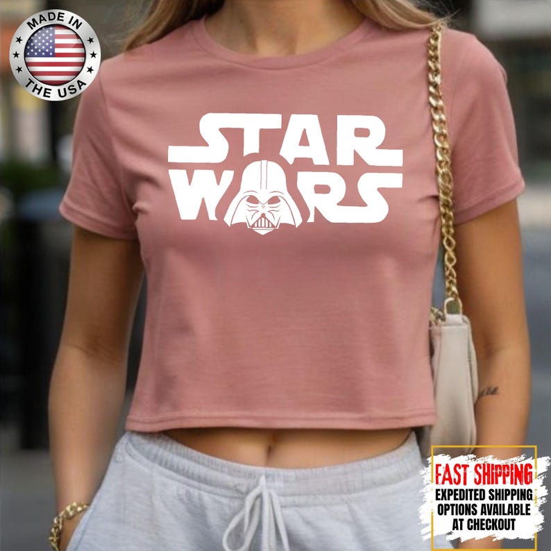 Puede incluir: Top corto rosa polvoriento con el texto blanco "STAR WARS" y un gr&aacute;fico de Darth Vader. Fabricado en EE. UU.