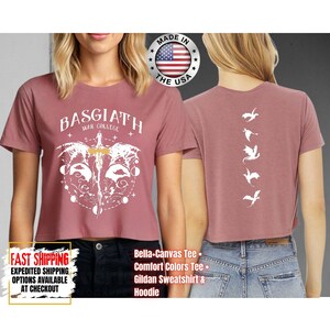 Puede incluir: Top corto rosa polvoriento con el texto "BASGIATH WAR COLLEGE" y un gr&aacute;fico blanco de una espada y alas. La parte posterior de la camisa presenta una fila vertical de siluetas de dragones blancos. Hecho en los EE. UU.