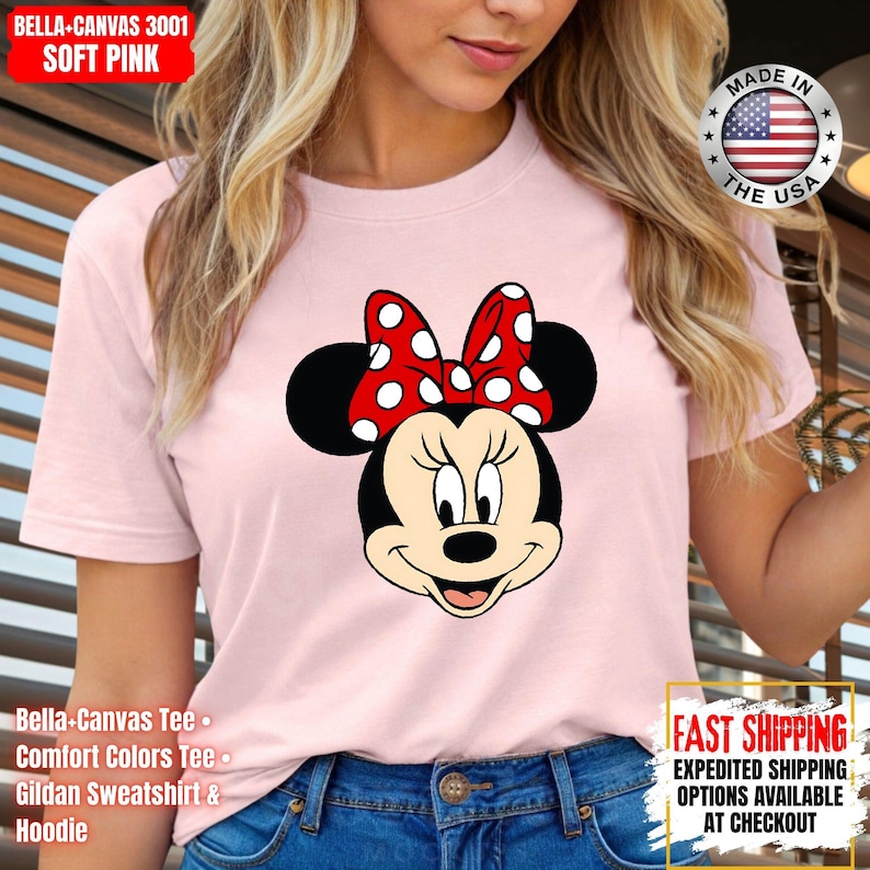 May include: A soft pink Bella+Canvas t-shirt featuring a Minnie Mouse graphic. The graphic shows Minnie's face with a red polka-dot bow. The shirt has the text "Bella-Canvas Tee", "Comfort Colors Tee", and "Gildan Sweatshirt & Hoodie".