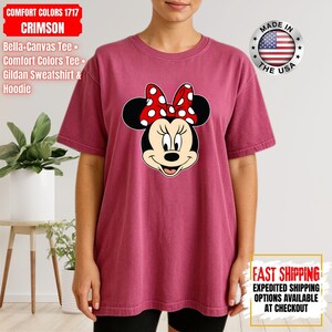 May include: Crimson Comfort Colors t-shirt featuring a Minnie Mouse graphic. The graphic shows Minnie's face with a red and white polka dot bow. The shirt has short sleeves and a relaxed fit. The text "COMFORT COLORS 1717 CRIMSON" is visible.