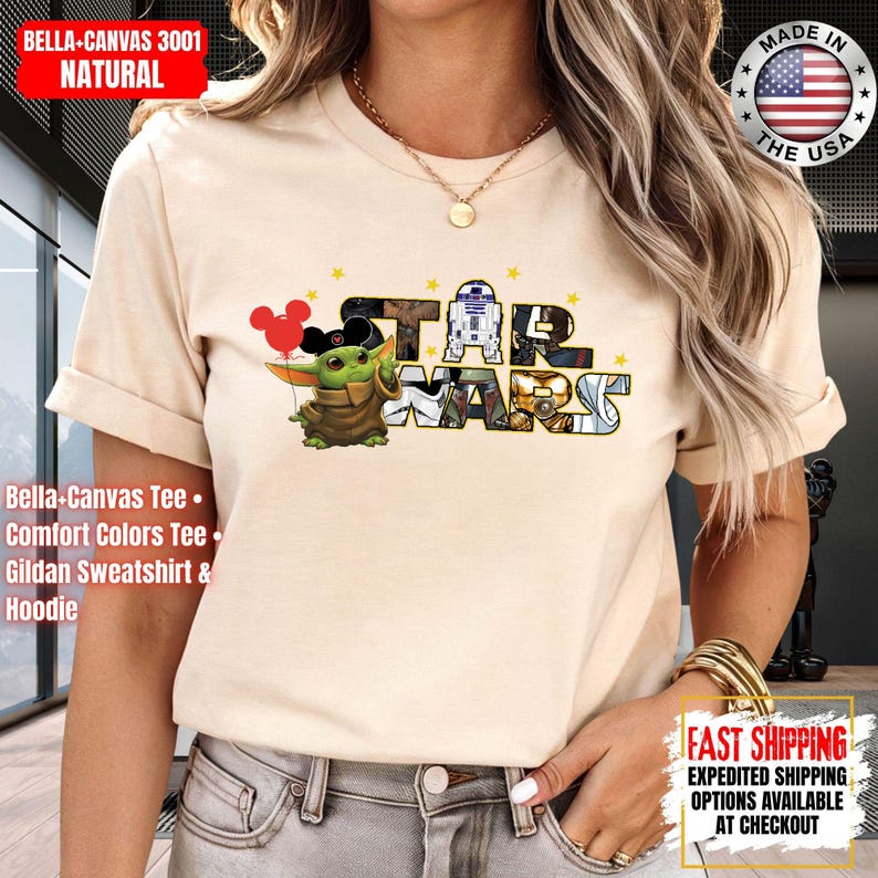 May include: A beige t-shirt featuring a Star Wars graphic with Baby Yoda, R2-D2, and the Star Wars logo. The shirt has short sleeves and is made of a soft material. The text "BELLA+CANVAS 3001 NATURAL" is in the upper left corner.