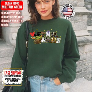 May include: A dark green Gildan sweatshirt featuring a Star Wars graphic with Baby Yoda and other characters. The text "STAR WARS" is displayed in a stylized font. The sweatshirt also has the text "GILDAN 18000 MILITARY GREEN".