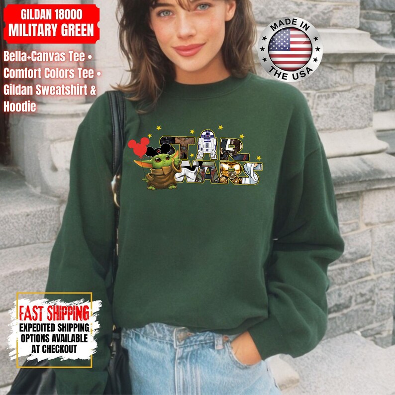May include: A dark green Gildan sweatshirt featuring a Star Wars graphic with Baby Yoda and other characters. The text "STAR WARS" is displayed in a stylized font. The sweatshirt also has the text "GILDAN 18000 MILITARY GREEN".