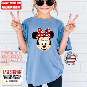 May include: A light blue t-shirt featuring a Minnie Mouse graphic with a red and white polka dot bow. The shirt has the text "COMFORT COLORS 9018 WASHED DENIM" at the top. The shirt is made in the USA.