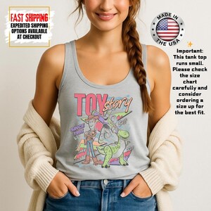 Op de afbeelding: Een lichtgrijze tanktop met de woorden "TOY Story" in roze en een afbeelding van Woody, Buzz Lightyear en Rex. De tekst "SINCE 1995" is ook zichtbaar. De tanktop heeft een ronde hals en is gemaakt in de VS.