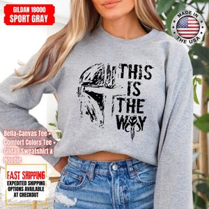 May include: Sport gray Gildan sweatshirt with a black graphic of a helmet and the text "THIS IS THE WAY". The sweatshirt is made in the USA. The image also includes the text "Bella-Canvas Tee, Comfort Colors Tee, Gildan Sweatshirt & Hoodie".