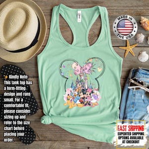 May include: Mint green tank top featuring a Minnie Mouse design with a castle, characters, and a pink bow. The tank top has a racerback style and is made in the USA. The image also includes a straw hat, sunglasses, and a pair of denim shorts.