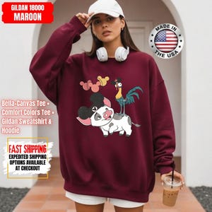 May include: Maroon Gildan sweatshirt featuring a cartoon illustration of a pig with Mickey Mouse ears, a rooster, and Mickey Mouse-shaped balloons. The text "MADE IN THE USA" is on the right. The sweatshirt also has the text "Bella+Canvas Tee", "Comfort Colors Tee", and "Gildan Sweatshirt & Hoodie".
