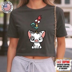 May include: Dark gray crop top featuring a cartoon illustration of a pig with a rooster on its head. The pig is white with black spots and pink ears. The rooster is red, yellow, and blue. The shirt has a "Made in the USA" graphic.
