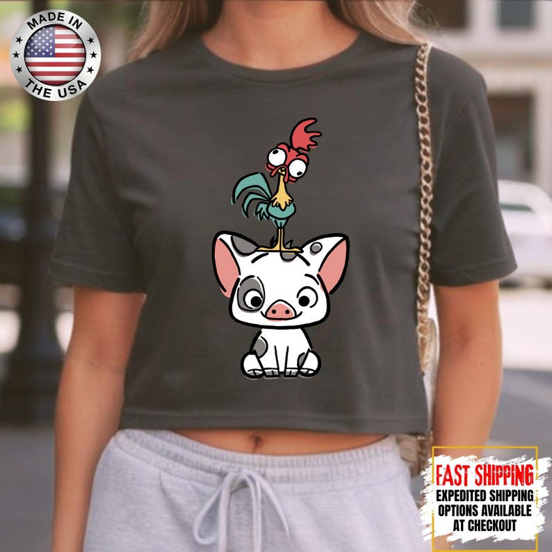 May include: Dark gray crop top featuring a cartoon illustration of a pig with a rooster on its head. The pig is white with black spots and pink ears. The rooster is red, yellow, and blue. The shirt has a "Made in the USA" graphic.
