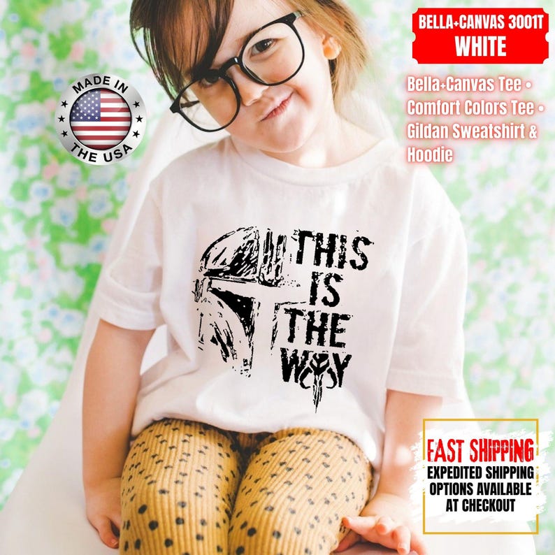 May include: White t-shirt with a black graphic of a helmet and the text "THIS IS THE WAY". The shirt is worn by a child wearing glasses. The image also includes text that says "MADE IN THE USA".