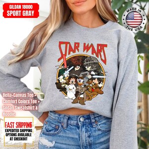 Op de afbeelding: Sportgrijs Gildan 18000 sweatshirt met een Star Wars-afbeelding met stripfiguren. Het ontwerp bevat de tekst "STAR WARS" in het rood. Het shirt heeft ook de tekst "Bella-Canvas Tee, Comfort Colors Tee, Gildan Sweatshirt & Hoodie".