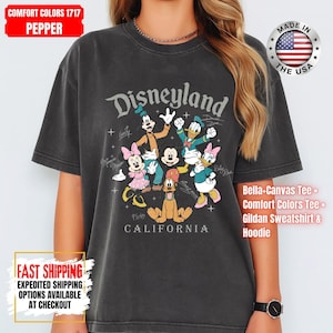 May include: A dark gray t-shirt featuring the word "Disneyland" above a graphic of Disney characters, including Mickey Mouse, Minnie Mouse, and Donald Duck. The shirt also has the text "CALIFORNIA" below the graphic.