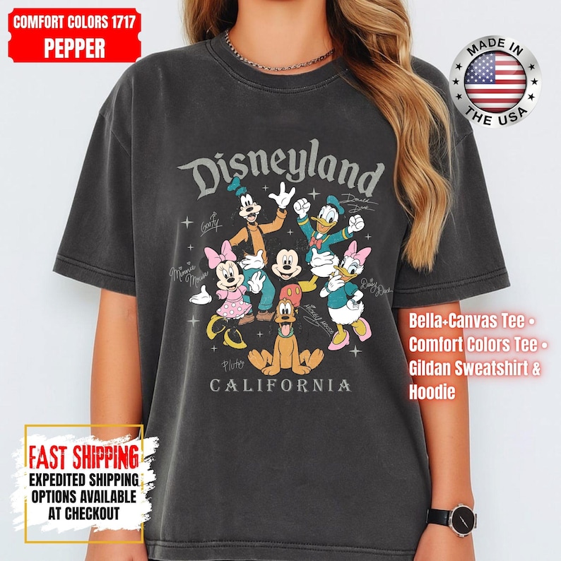 May include: A dark gray t-shirt featuring the word "Disneyland" above a graphic of Disney characters, including Mickey Mouse, Minnie Mouse, and Donald Duck. The shirt also has the text "CALIFORNIA" below the graphic.