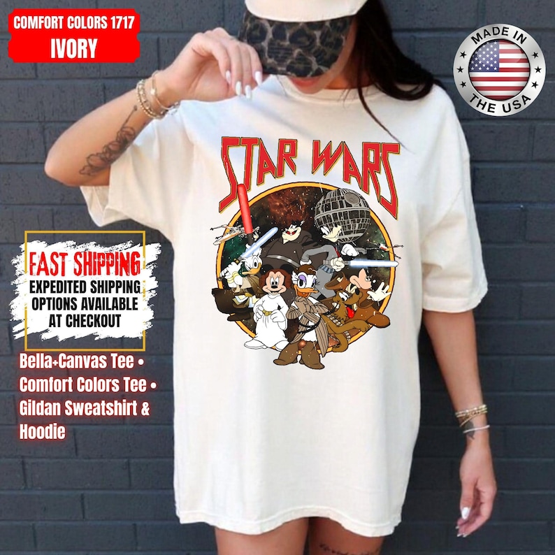Op de afbeelding: Ivoor kleurig Star Wars grafisch T-shirt met een cartoon mix van Star Wars en Disney karakters. Het ontwerp bevat het Star Wars logo in rood, een Death Star en diverse karakters. Het shirt is een Comfort Colors 1717 Bella-Canvas Tee.